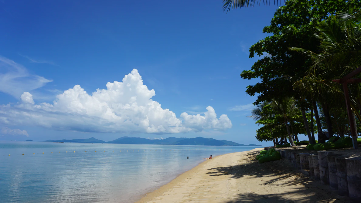 An uncrowded beach in Koh Samui. There is blue water and golden sand, and there are mountains in the distance.