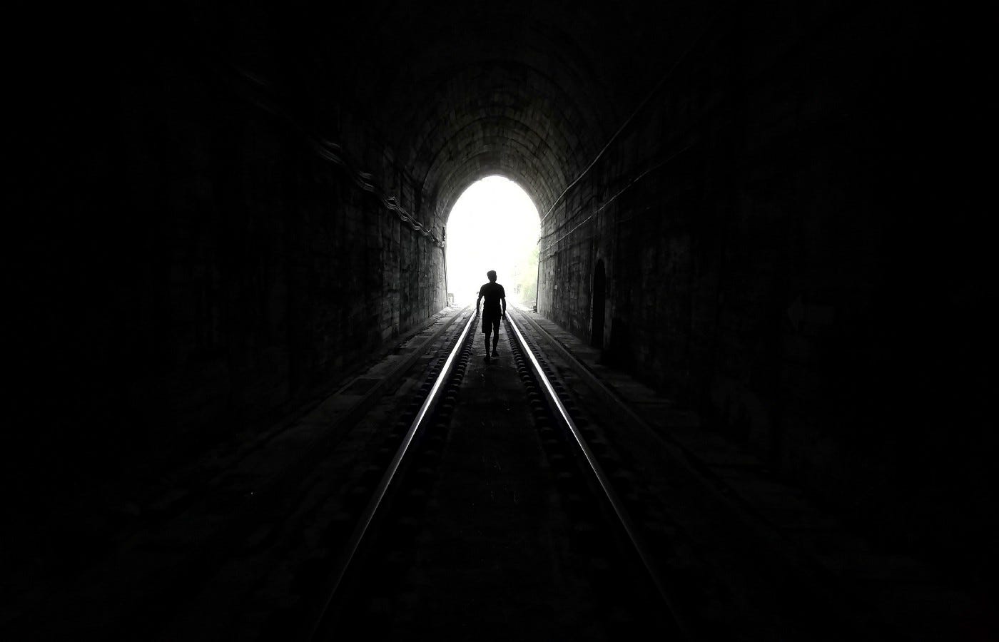 Man waking between train tracks toward the light at the end of a train tunnel.