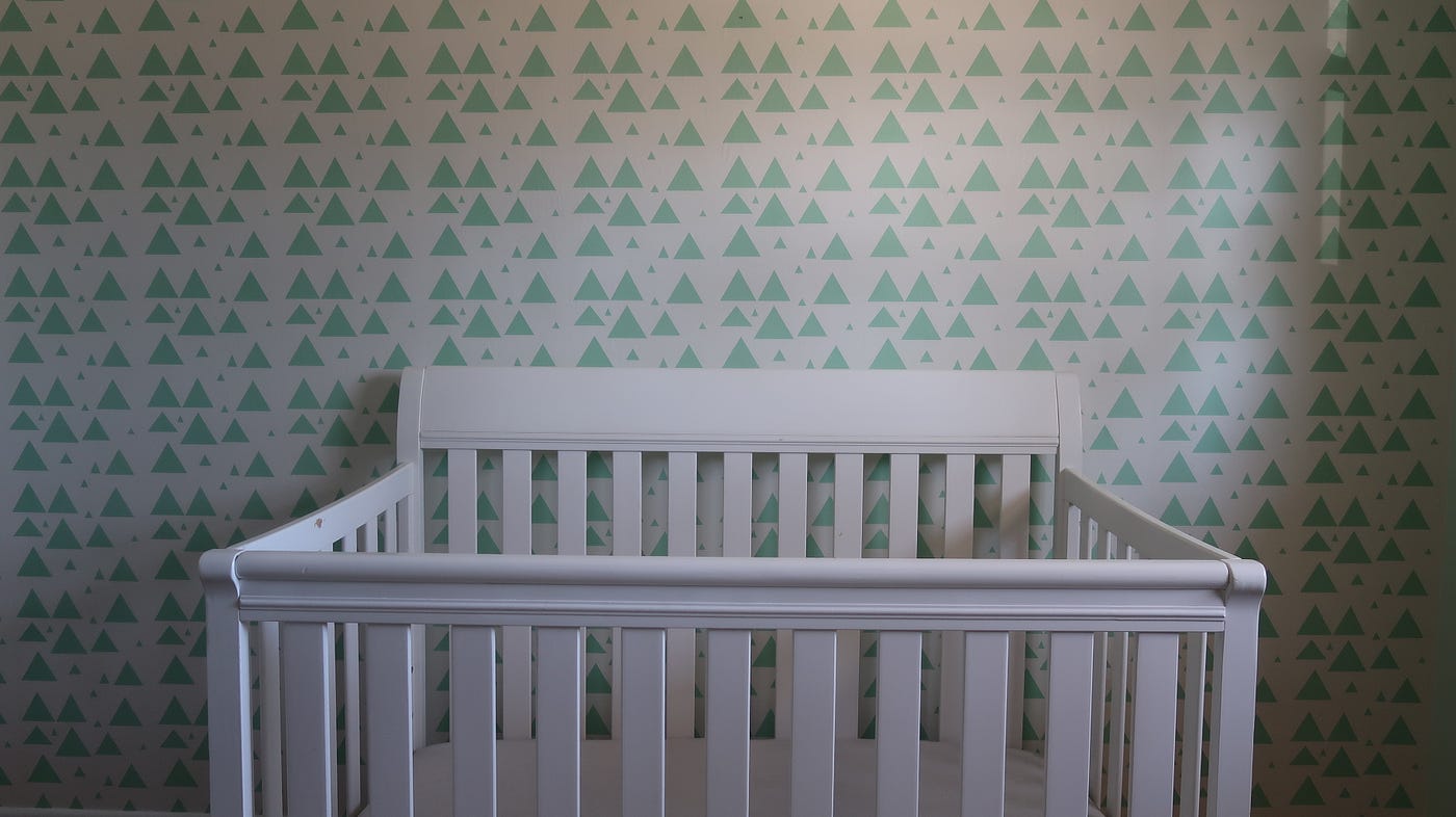 A white crib against a green triangle wallpapered wall.