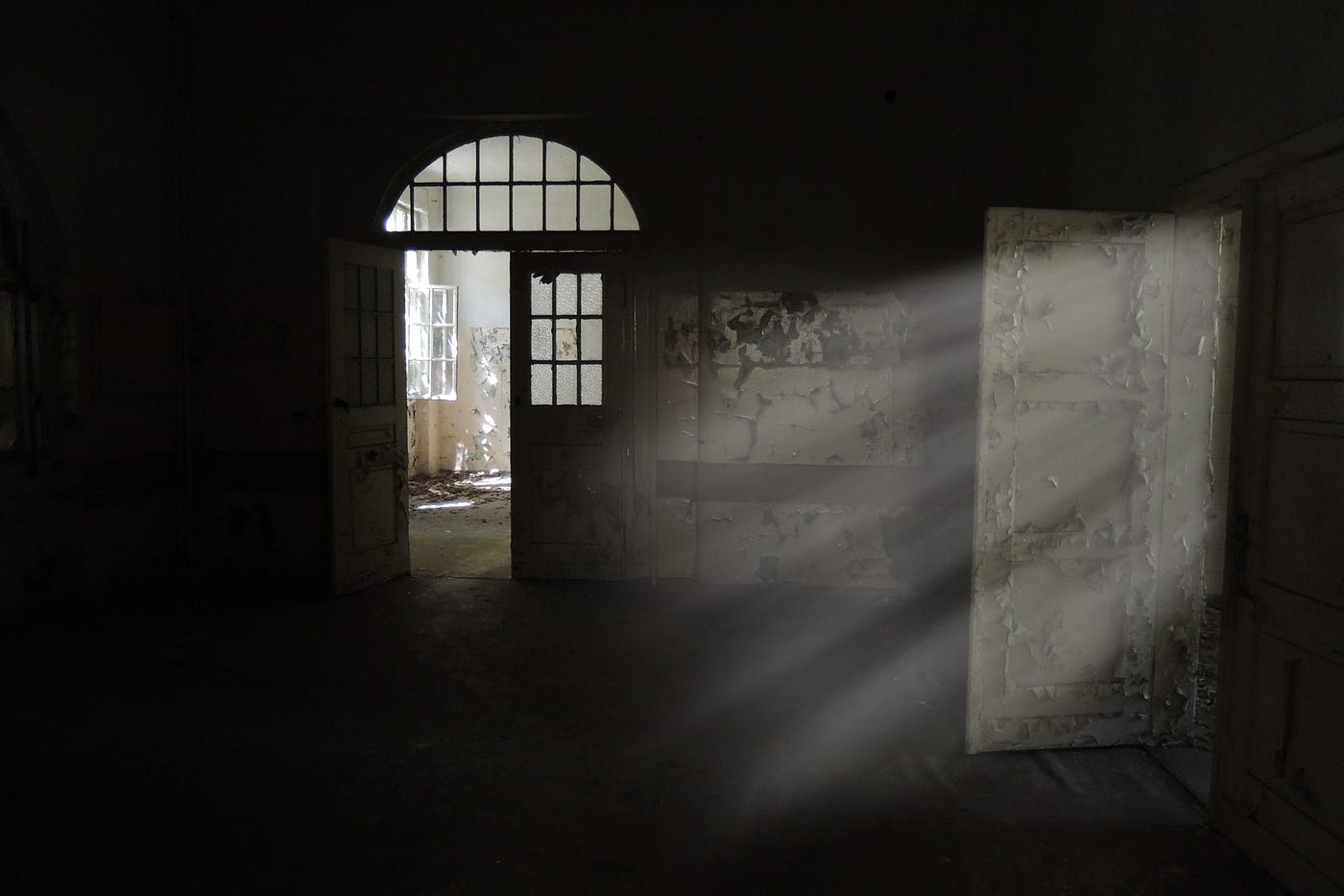 A dark, deserted room is lit up by shafts of sunlight through a doorway. The walls are peeling, and through another door we can see a similar room, also lit by dappled daylight.