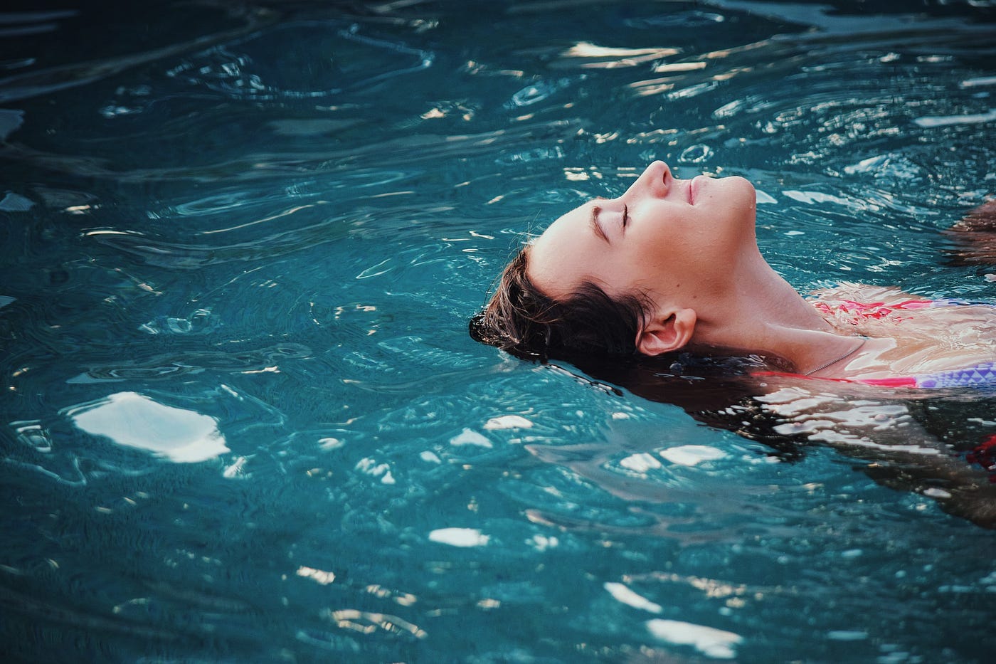A woman floats in a pool. We see her from upper chest level up, emerging from the right side of the image. She is white and has brown hair.
