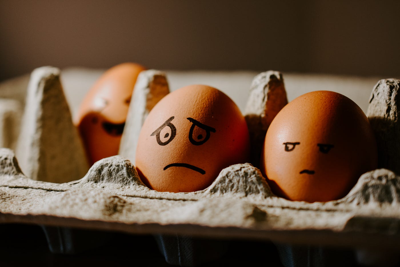 Three brown eggs in a cardboard carton, two in the front and one in the back. Someone has drawn sad faces on the front ones.