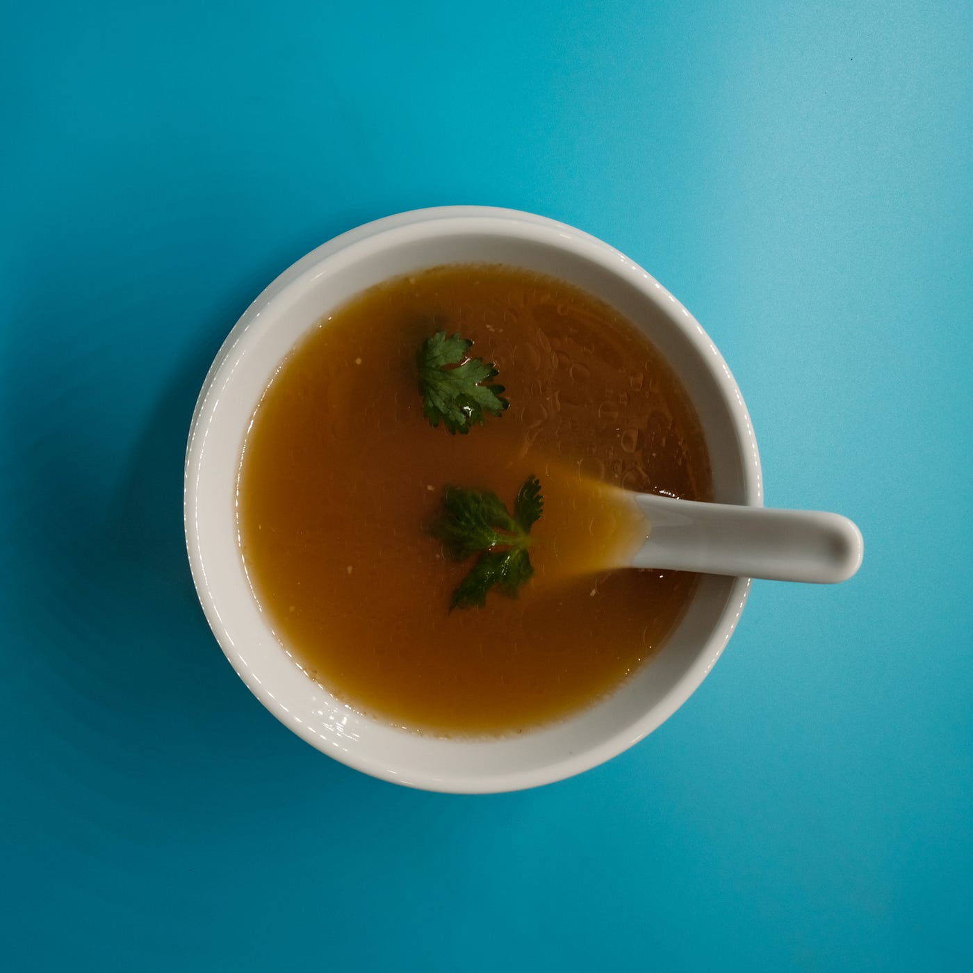 A bowl of miso, as seen from above. The bowl is white, and a white spoon is perched in the brownish soup. Light blue table.