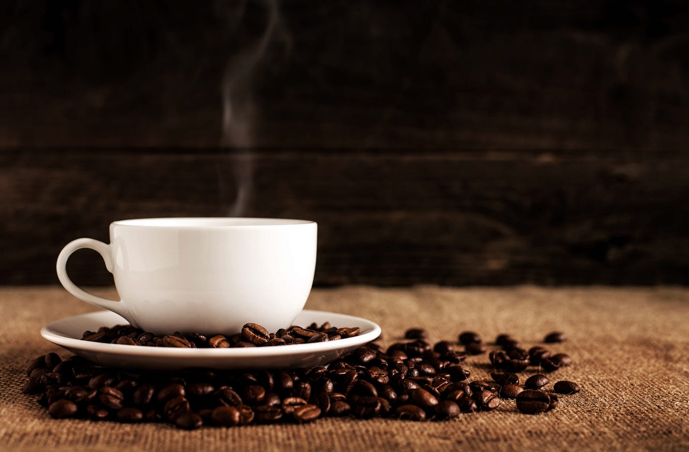 A white cup (with a small curl of steam rising from it) sits on a white saucer, with numerous coffee beans scattered in the saucer and outside of it. A new study suggests that coffee extends life, even if we add sugar.