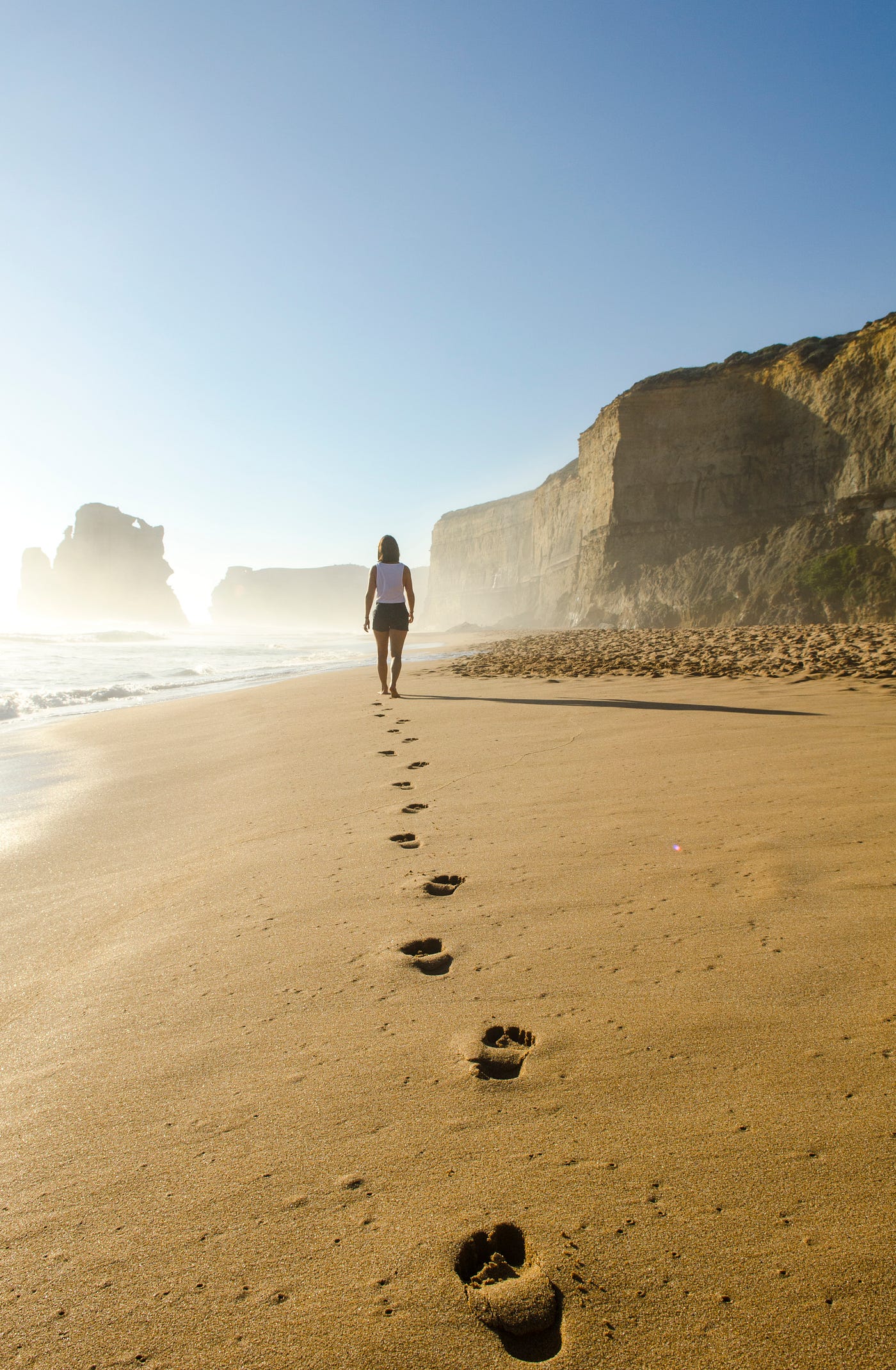 A woman, wearing a white T-shirt and dark blue shorts, is at a moderate distance from us, on a sandy beach. She walks away from us, leaving her footprints in the sand. Walking 10,000 steps a day can help you achieve several health benefits, such as improved cardiovascular health, increased energy levels, and weight management.