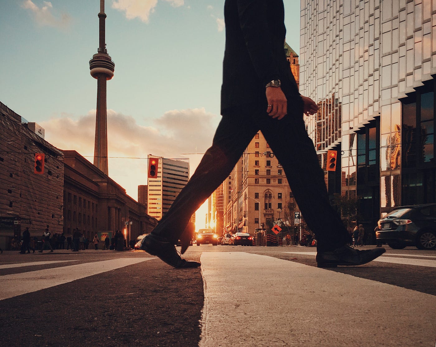 A man walks in Toronto, with the CN Tower in the background. He is in a crosswalk and in business attire. We see him from chest level down, stride wide open. The sun rises (or sets) in the background.
