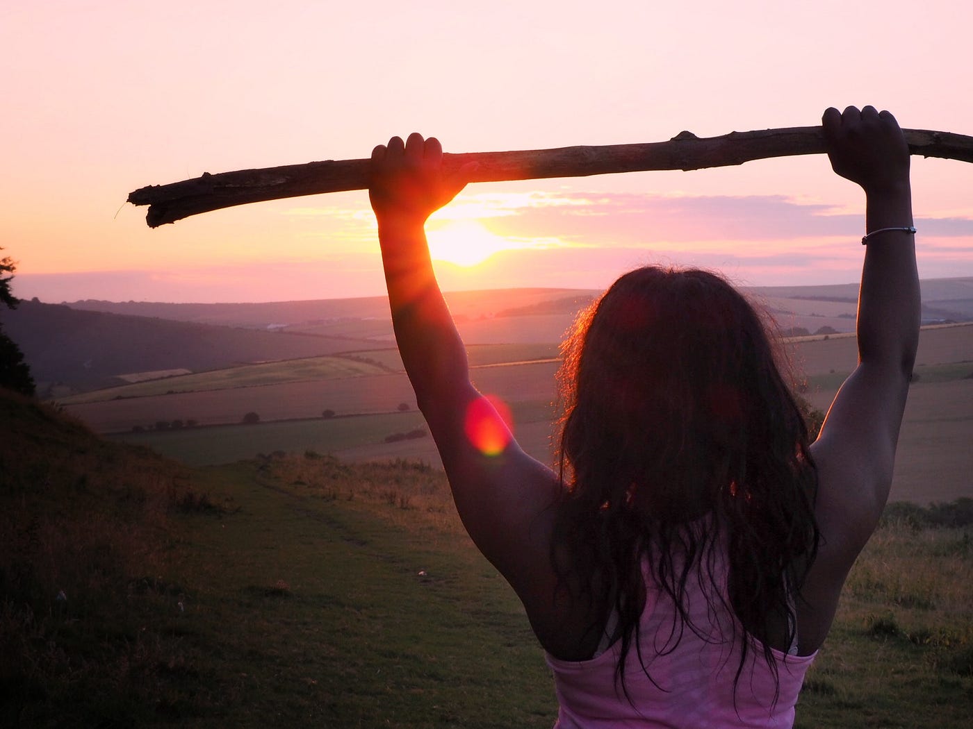 Girl at sunset holding a stick over her head, backlit by the sun.