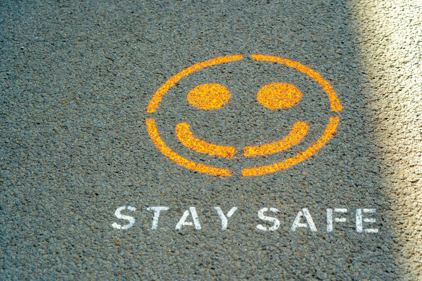 Spray painted on gray pavement are the words “STAY SAFE” in capital letters, colored white. Just above the words, we see a yellow smiley face.