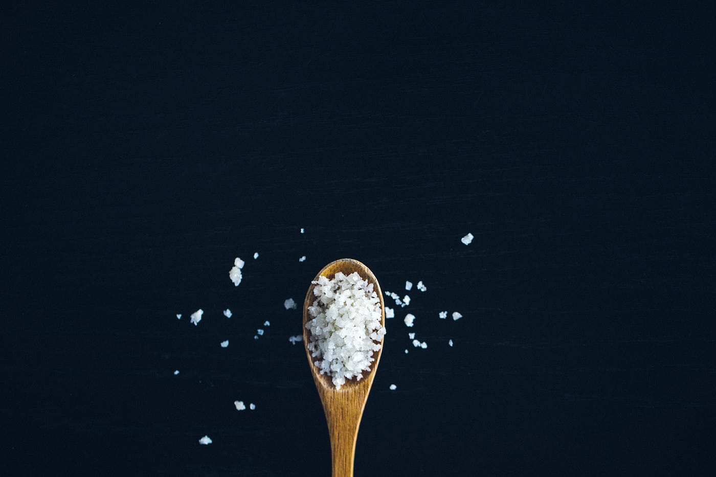 A small wooden spoon emerges from the bottom of the image, holding coarse salt that is spilling onto the black background. Limiting sodium intake is one of the 5 natural ways to drop your blood pressure.