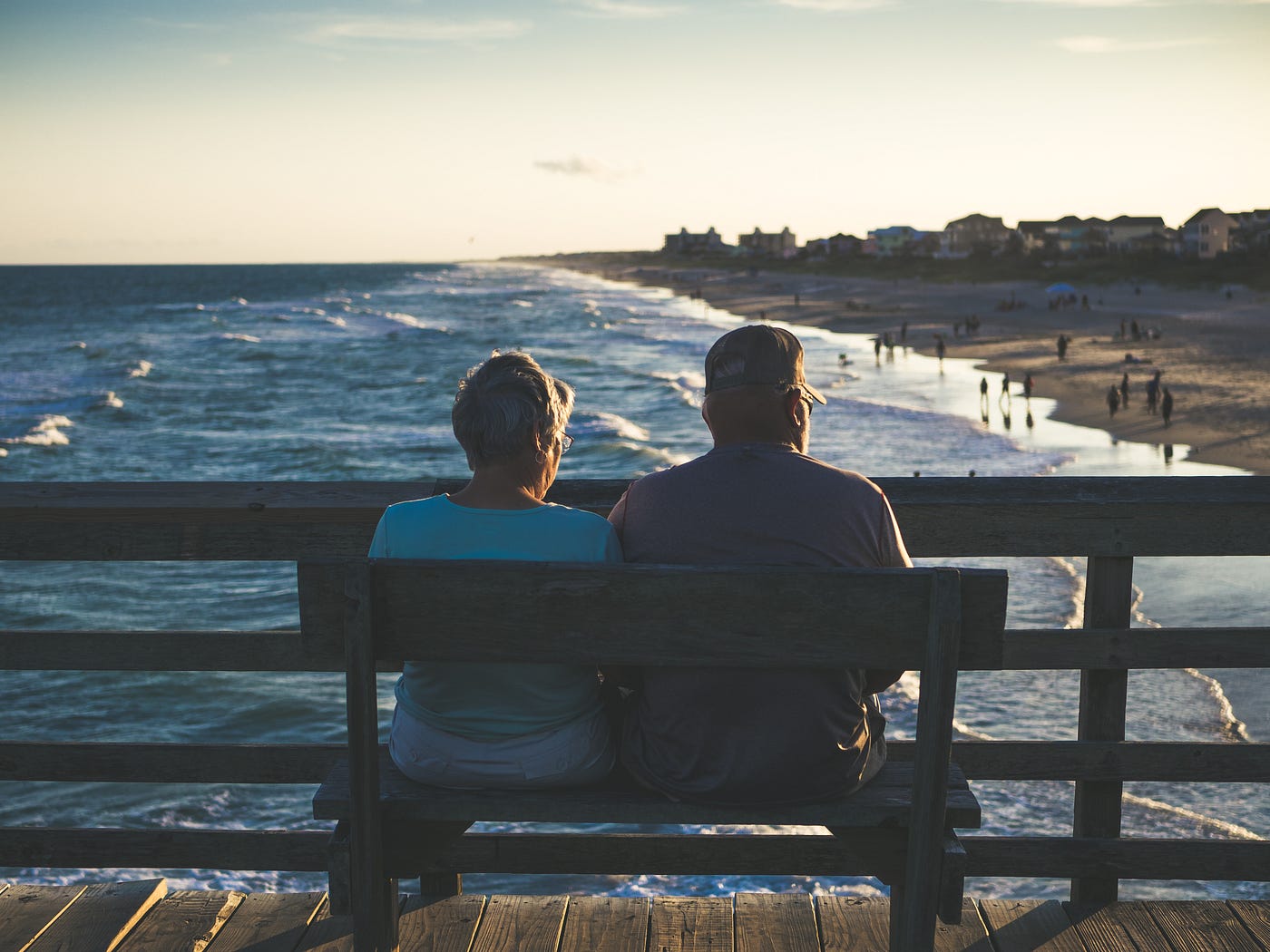 An older couple sits on a bench, facing away from us as they look down at the ocean as it meats the beach.
