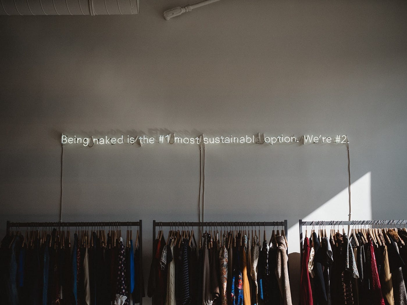 Neon lights above a clothing rack that reads: being naked is the #1 most sustainable option. we’re #2.