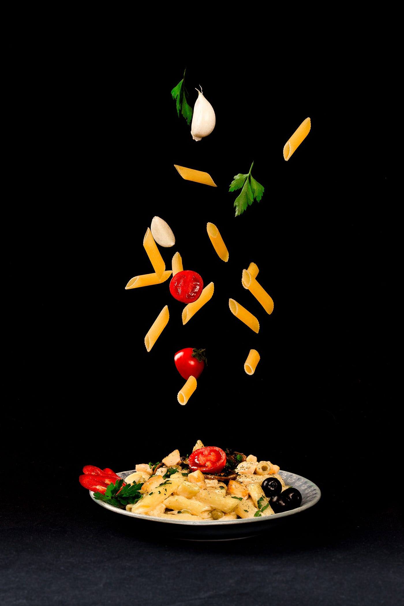 A plate of pasta and vegetables (including sliced tomatoes), with both also flying upward. Black background.