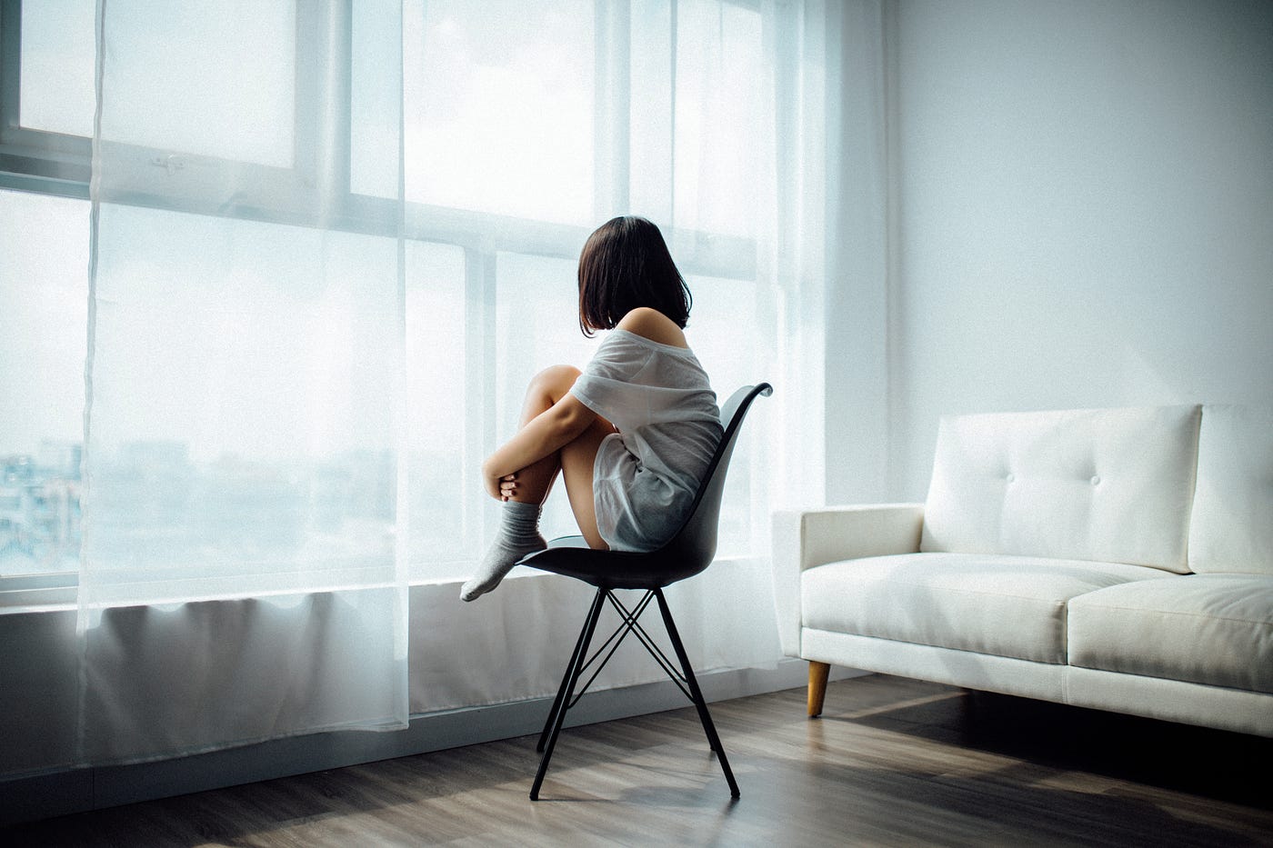 Person sitting on a chair with their knees to their chest, staring out a wide window.