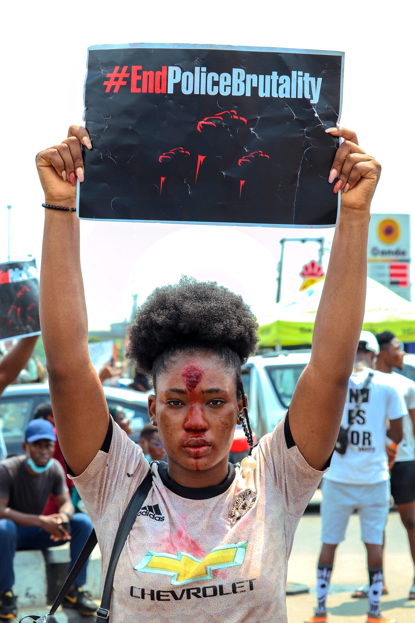 Attractive young woman holds aloft a sign saying, “EndPoliceBrutality” during a protest. She is costumed like a victim.