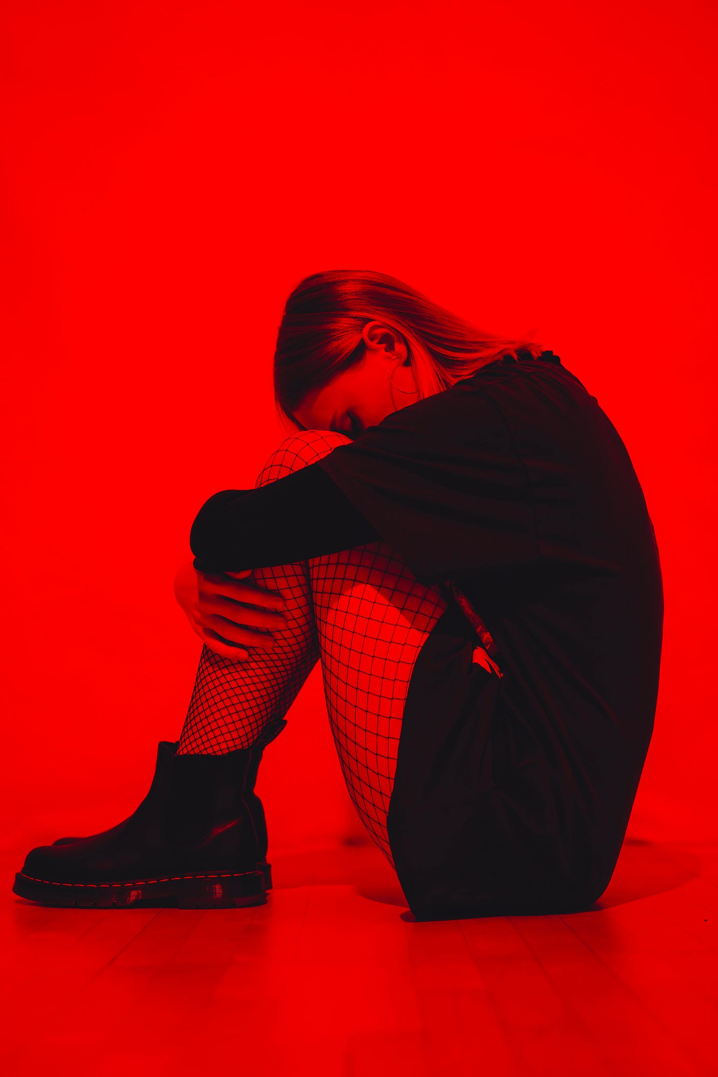 A depressed-appearing young woman sits, her head down while she embraces her bent lefts. Red background.
