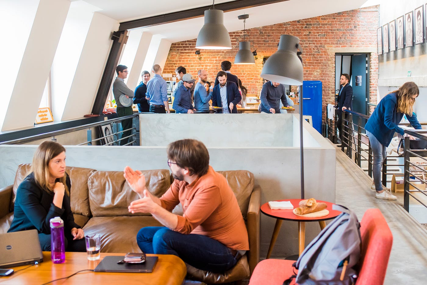 Photo of a bright, open-style office with two people conversing on a couch in the foreground and multiple people standing and conversing in the background.