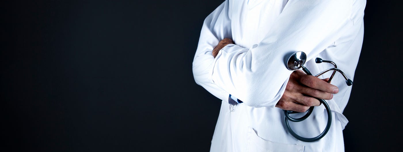 A doctor in three-quarter pose stands on the right side of the screen, arms folded with the right hand holding a curled stethoscope. Black background. We see the doctor from waist level to upper chest, wearing a white doctor’s jacket.