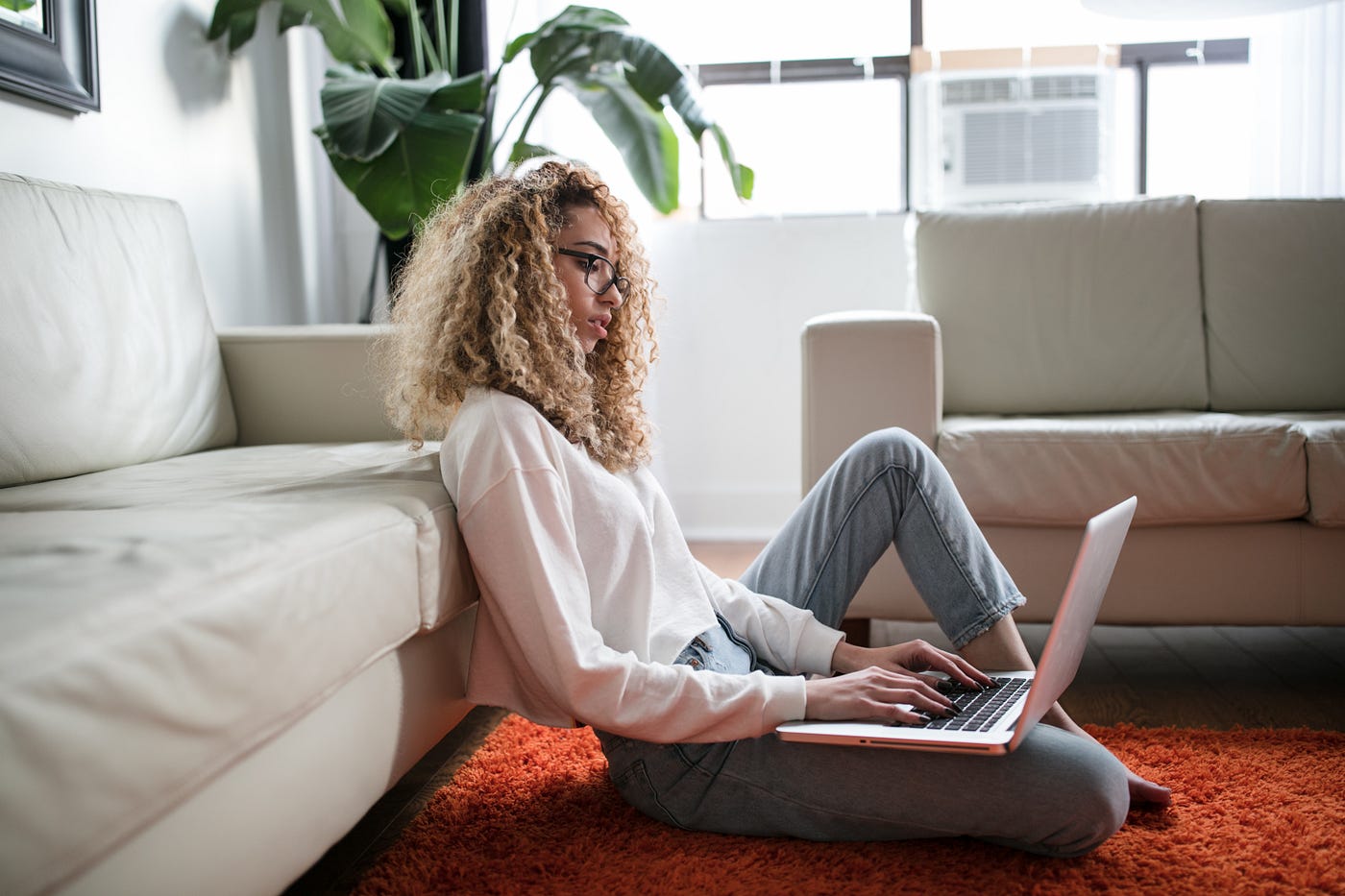 A woman sits on the floor leaning against a couch, and uses her laptop.