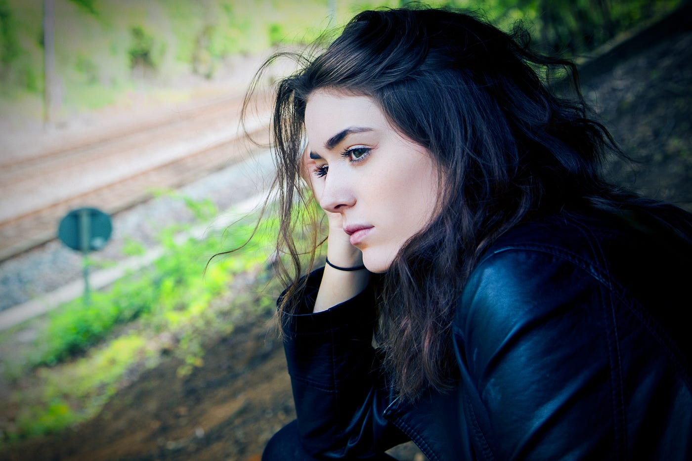 Young brunette woman seen in profile. Her right hand is on the side of her head as she looks to the left of the image. The woman looks pensive. Railroad tracks in the distance.