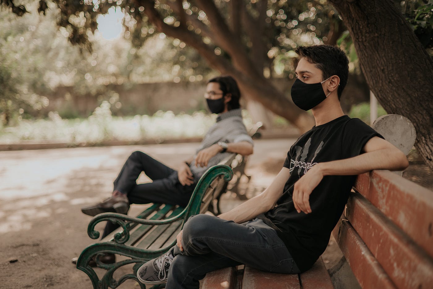 Two people sitting on park benches, seated far away from each other, wearing black cloth face masks