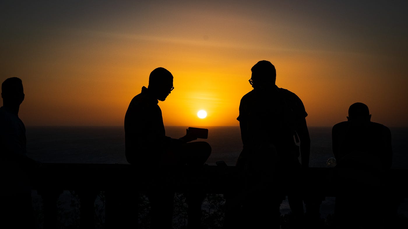Four men in shadows in the dark in front of a setting sun.