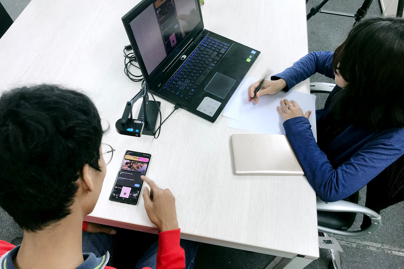 Two people in an office. One is testing a product on a phone, and the other is taking notes.