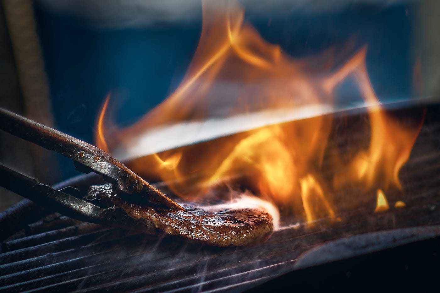 Tongs extend from the left to place a hamburger on a grill. Flames extend upward to the right of the meat.