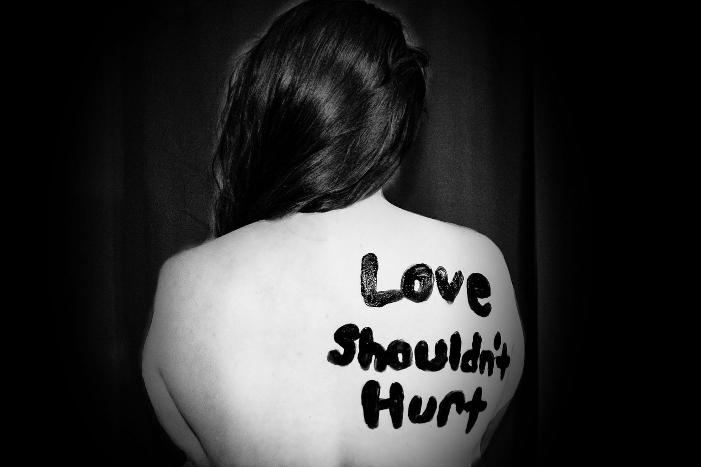 A naked girl with long dark hair is sat in darkness. ‘Love shouldn’t hurt’ is written on her back.