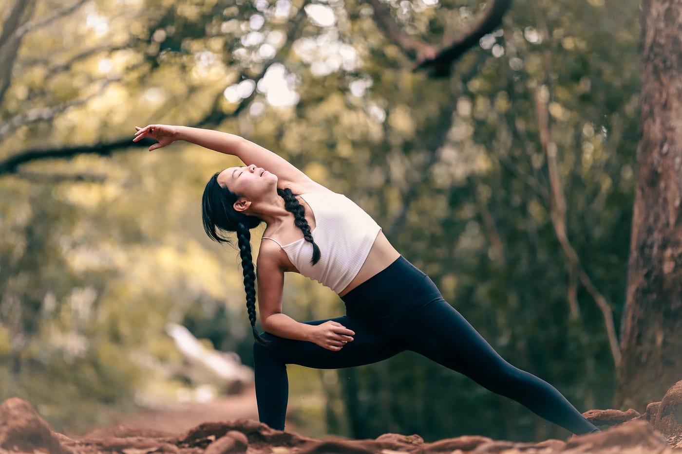 Woman stretches to her right, arm over her head, left leg extended and right leg bent. She wears a white shirt and black sweatpants.