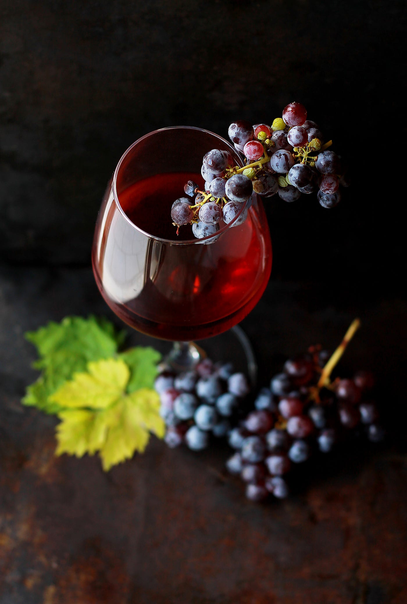 A glass (with a stem) is half filled with red wine. Purple grapes perch on the edge. Another bunch of grapes is on the table next to the glass.