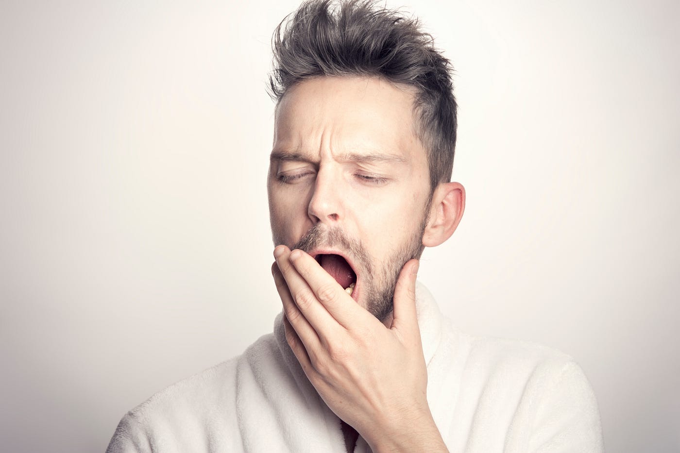 Young white male faces us, eyes closed as he yawns. He puts his left hand up to his mouth. White sweater, white background.