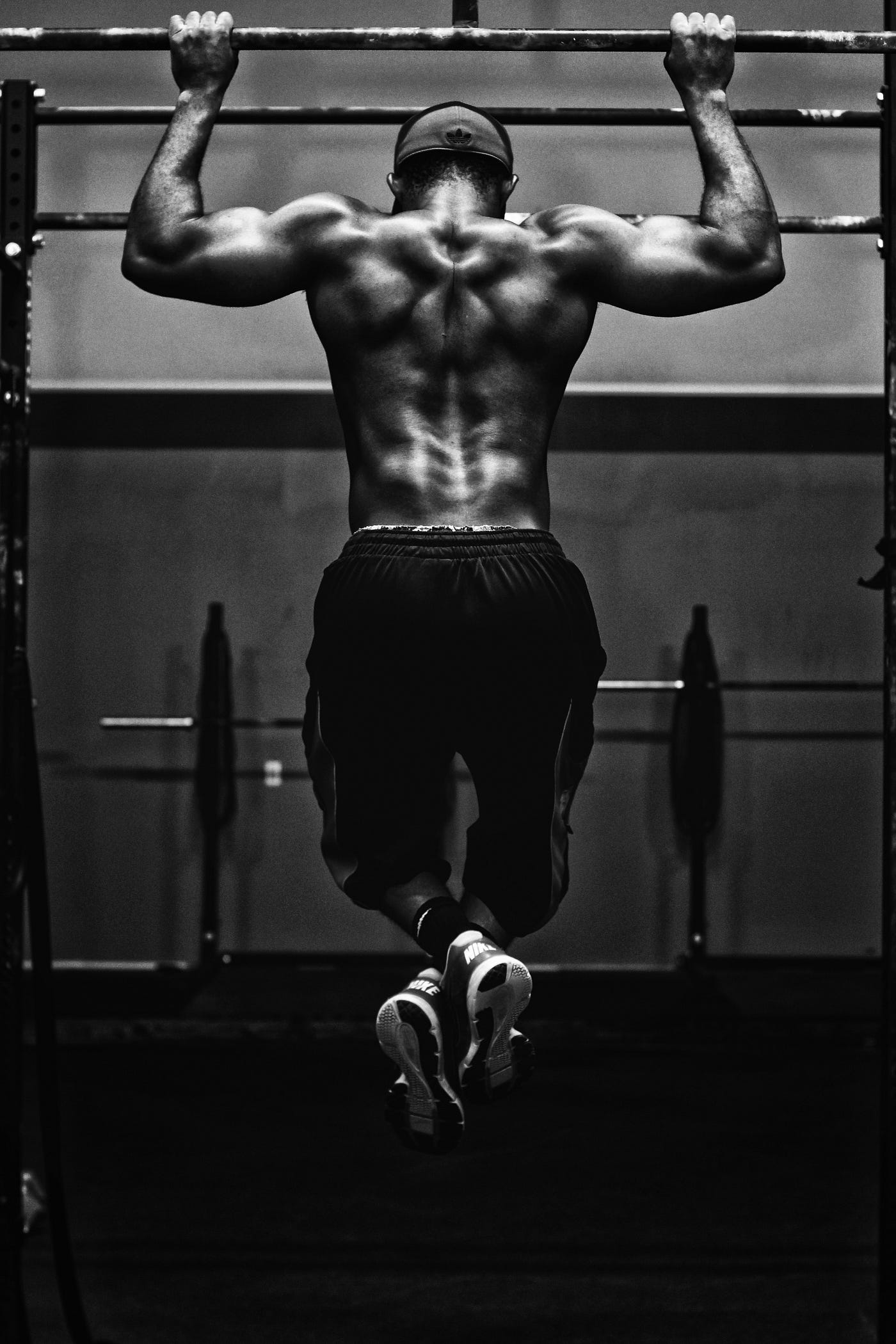 Black man doing a wide grip pull-up. We see him from behind, shirt off, muscles rippling. Black and white photo.