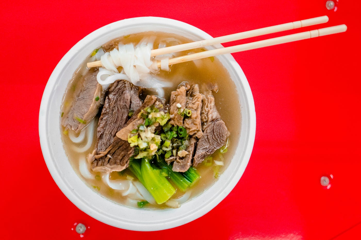 White bowl with meat, vegetables, and noodles. Two chopsticks are perched on the bowl at the two o’clock position. Red background.