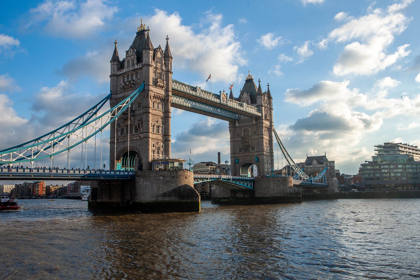 London’s Tower Bridge in daytime. Blue sky with some cumulus clouds. We see the bridge from a low perspective from across the river, diagonally.