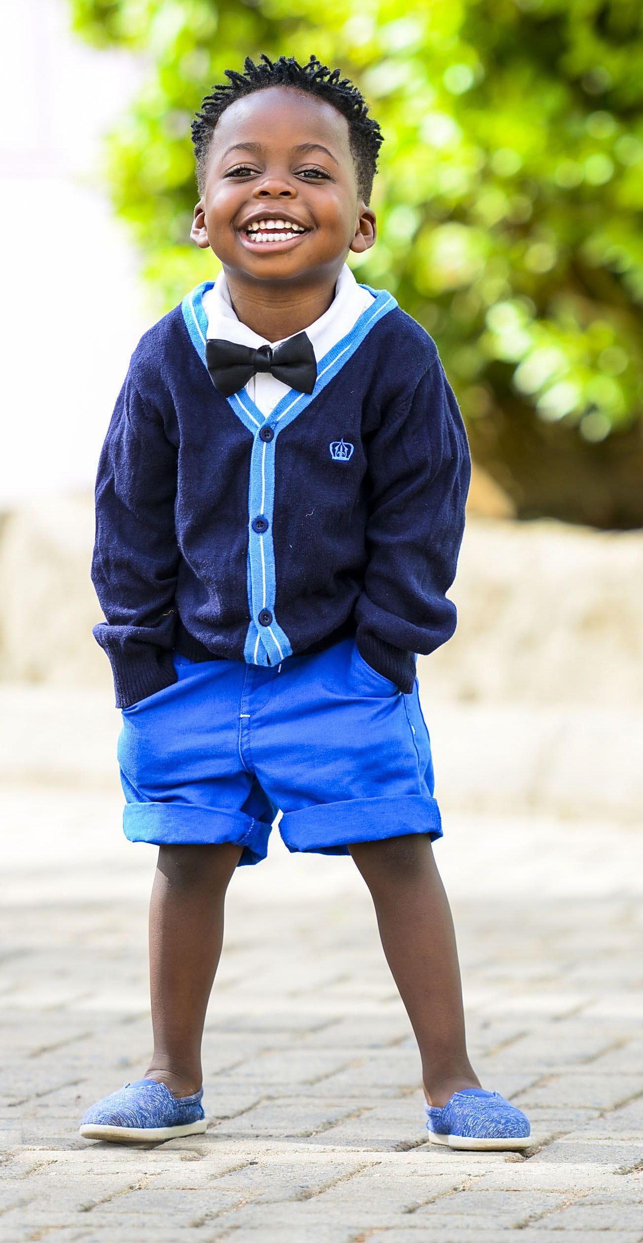 A young (around 4) black child, in a blue button down sweater and blue shorts, faces us as he smiles. His hands are in his pockets.