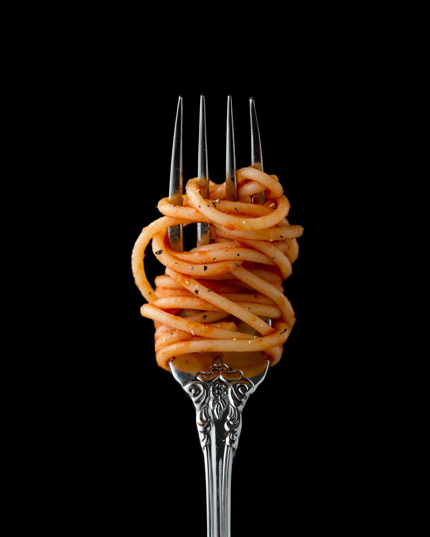 We see the end of a fork, pointing up from below, with spaghetti twirling around it. Black background.