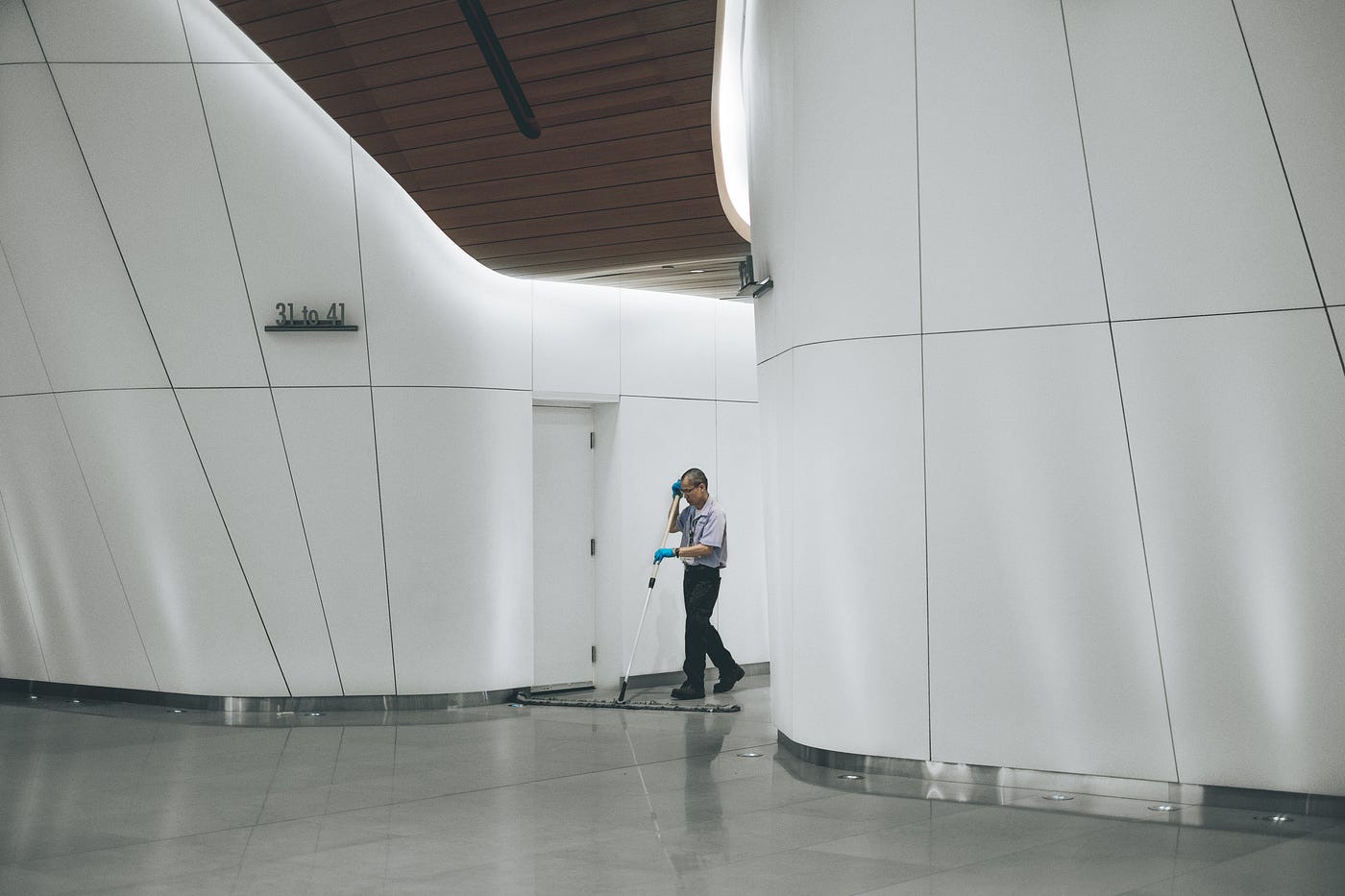 An Asian man sweeps with a long broom. He walks between white curing walls. The scene appears to be lit with fluorescent lights.
