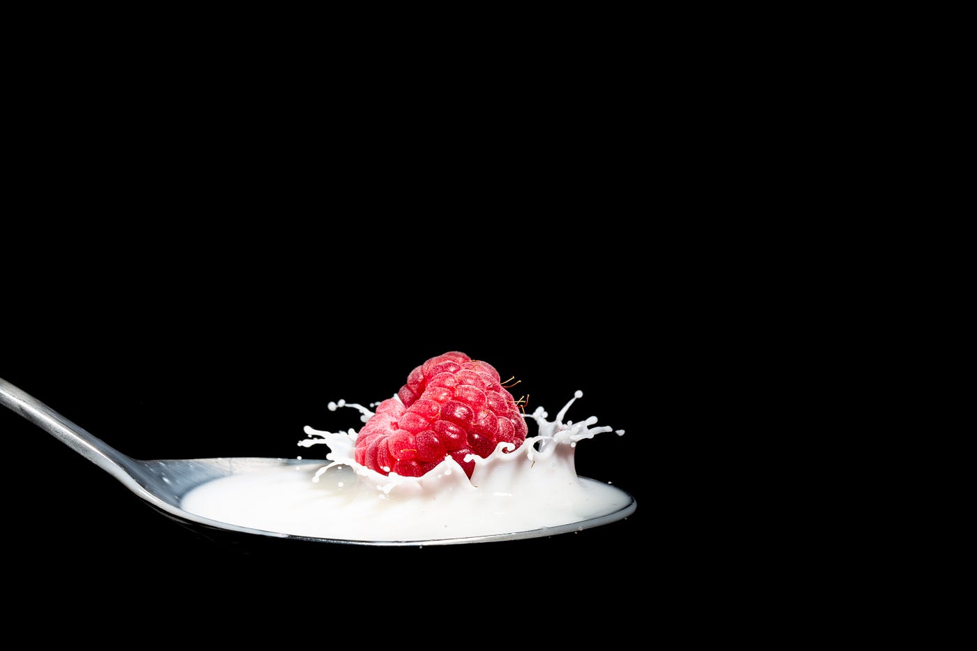A spoon emerges from the lower left of the screen, holding milk/yogurt with a raspberry splashing into it.