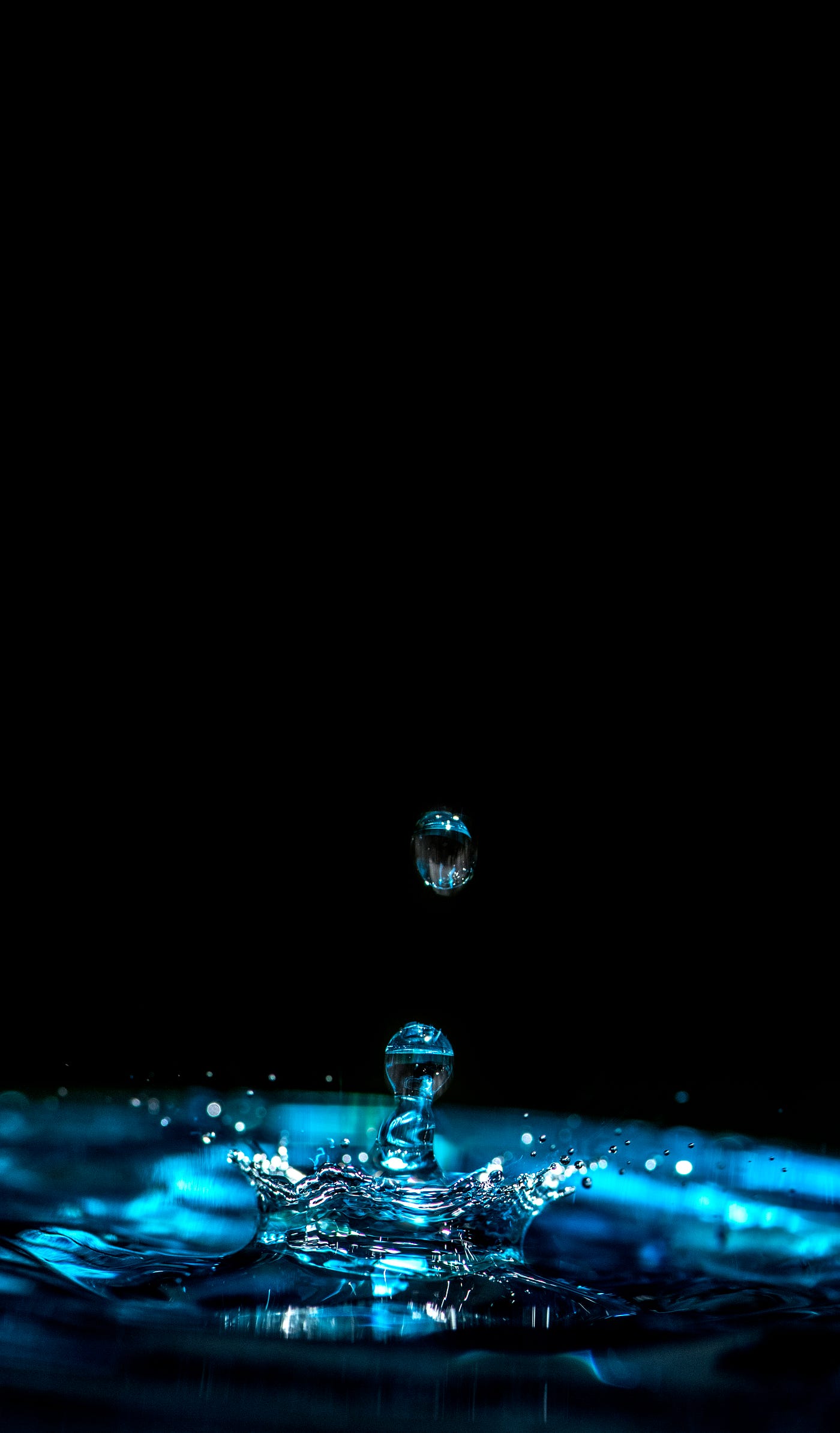 A photograph of a drop of water hitting a pool of water and bouncing up off of it. Black background.