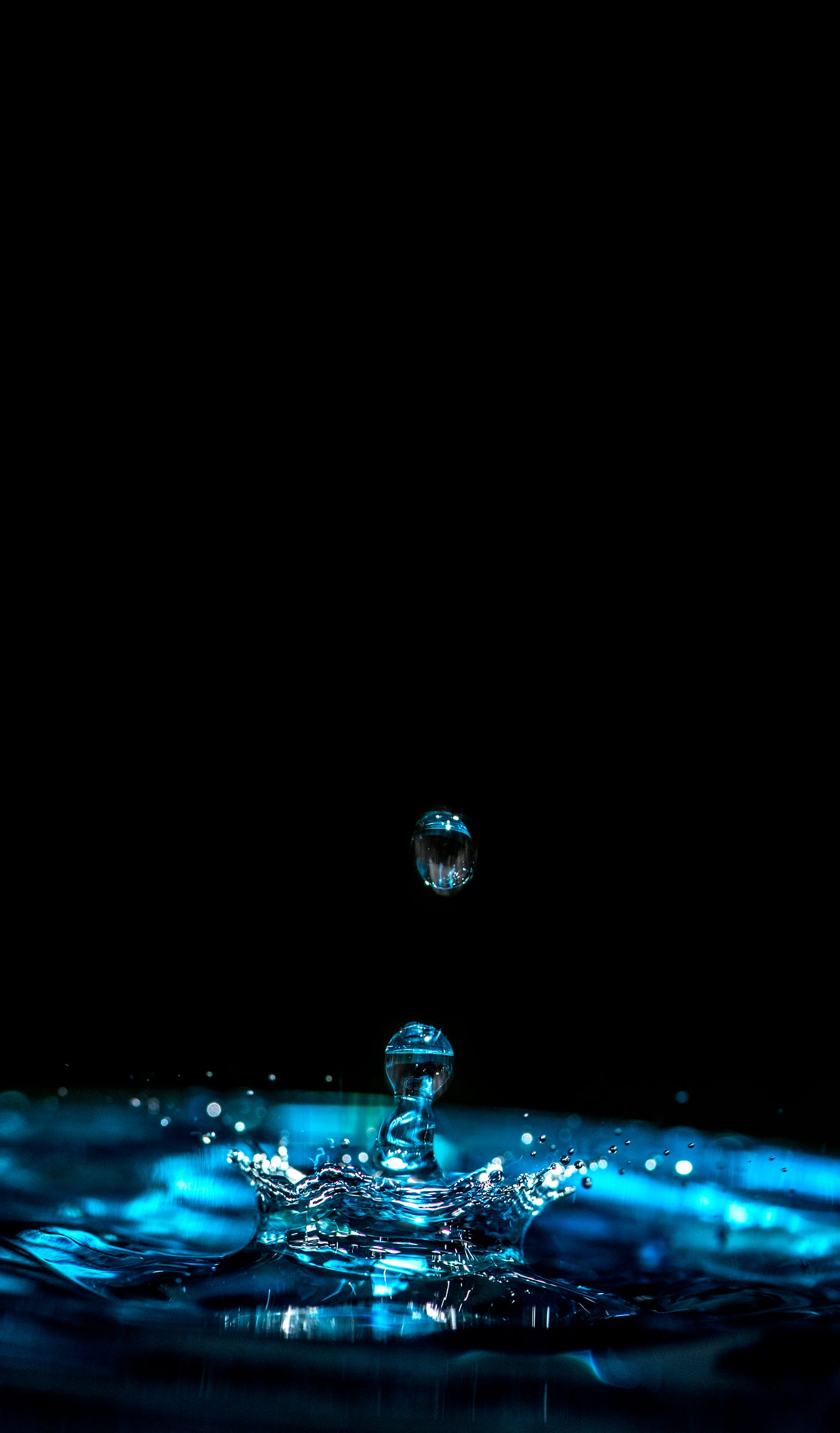 A photograph of a drop of water hitting a pool of water and bouncing up off of it. Black background.