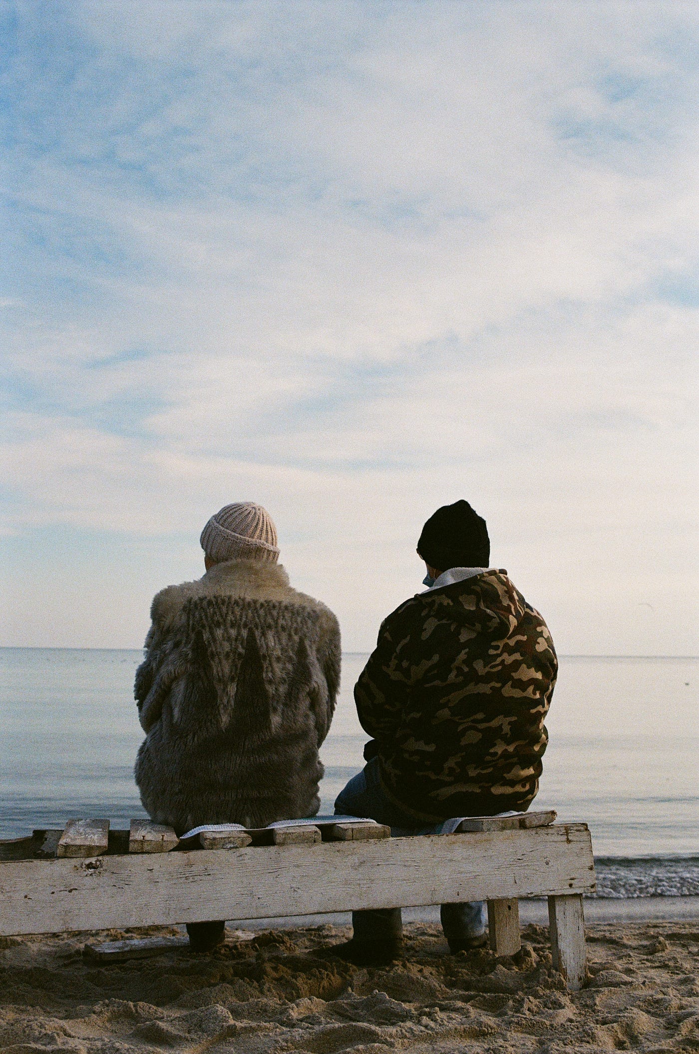 An older couple, dressed for winter with caps and patterned winter coats, sits facing away from us. They gaze over what looks like fog or clouds.