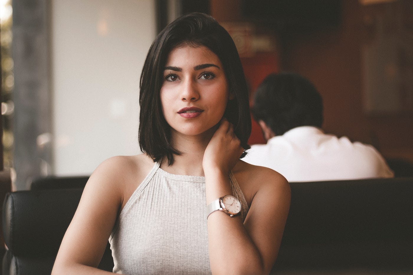 Young brunette woman, in her 30s, looks directly at the camera. A man is seated to the right in the distance, facing away. The woman wears a sleeveless white top.