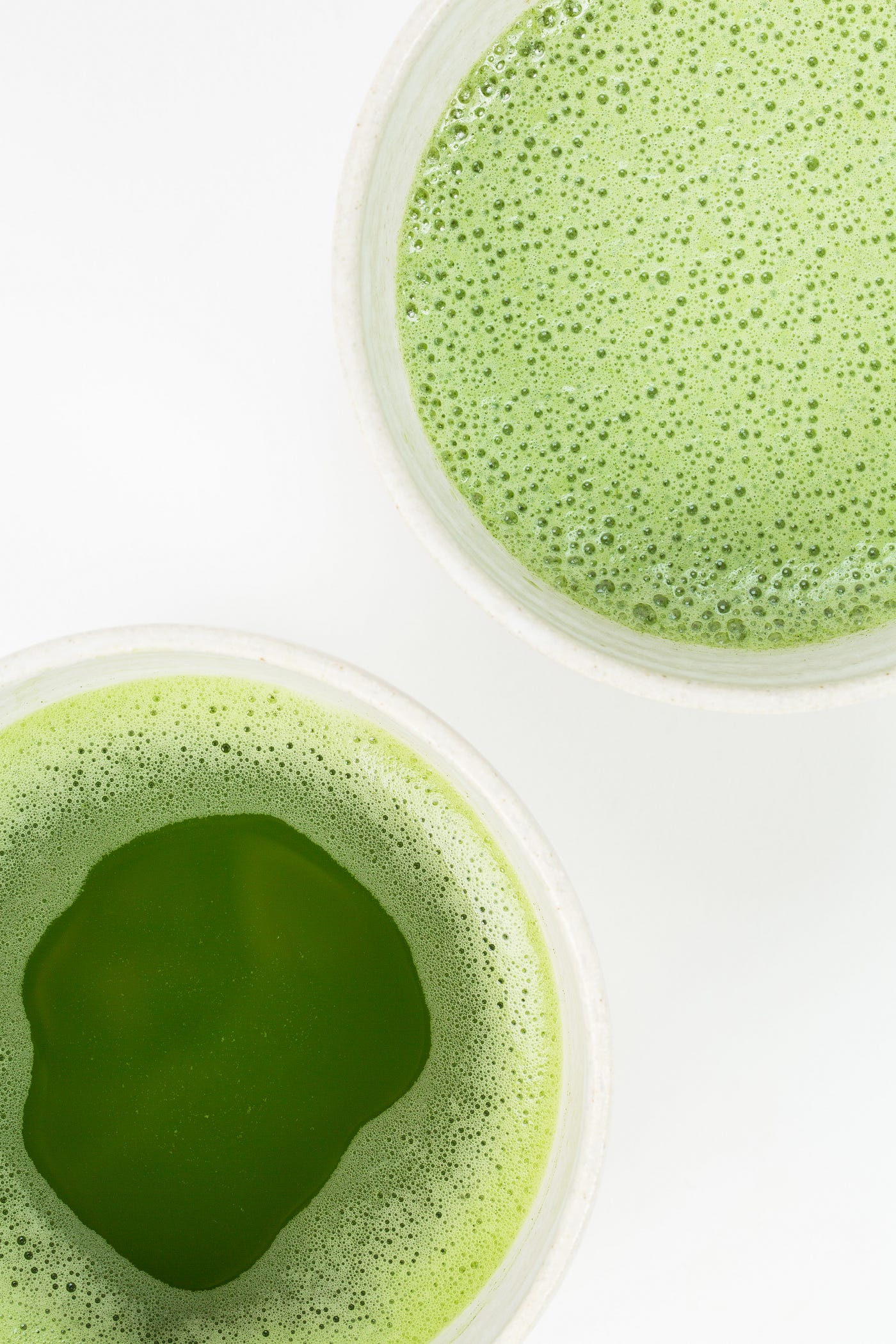 Two cups of green tea, slightly foamy (tiny bubbles). One sits in the upper right of the image, the other in the lower left. We see the cups from above. White background.