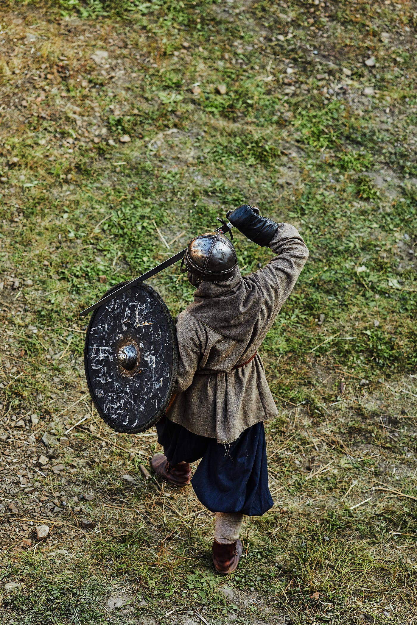 A medieval/Viking cosplayer is seen from above, standing alone in a muddy, grassy field. No face is visible. The individual’s back is to the viewer. They wear a metal helmet without horns, carry a dull metallic shield and hold their dull metal sword across their chest. They are dressed in a brownish-grey tunic top with a hood, the material is coarse. Their pants are billowed and navy blue. Their boots are brown leather and with canvas uppers.