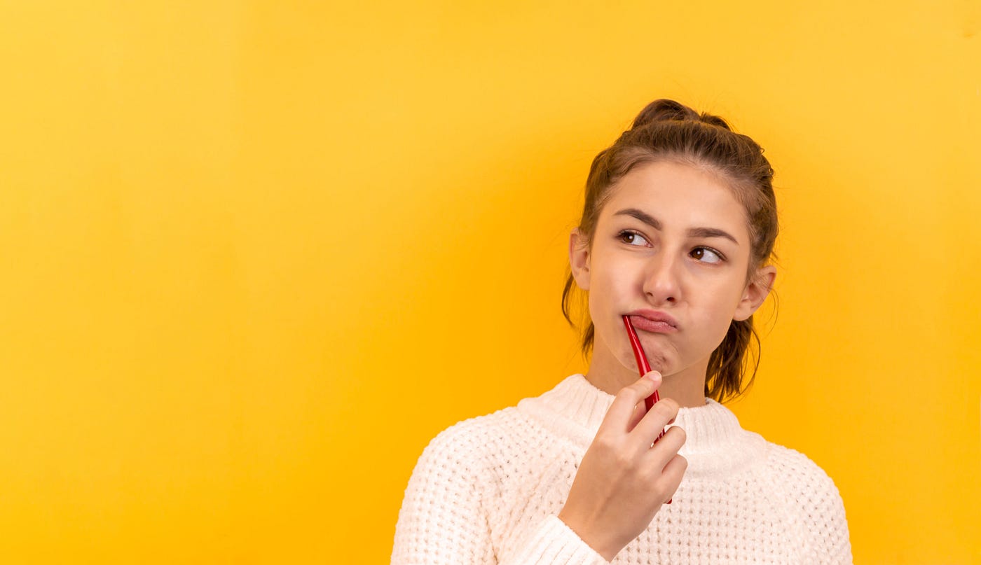 Young woman (in her late teens) holds a red toothbrush in the right side of her mouth. She wears a white turtle neck sweater. Yellow background.