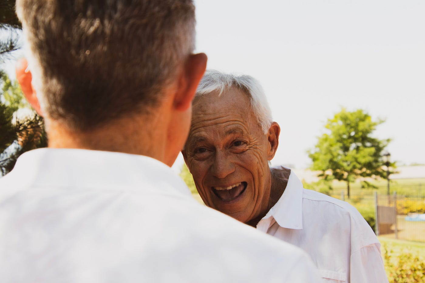 Older white man laughs. He wears a casual collared white shirt. He faces a man, the latter with his back to us in closeup.
