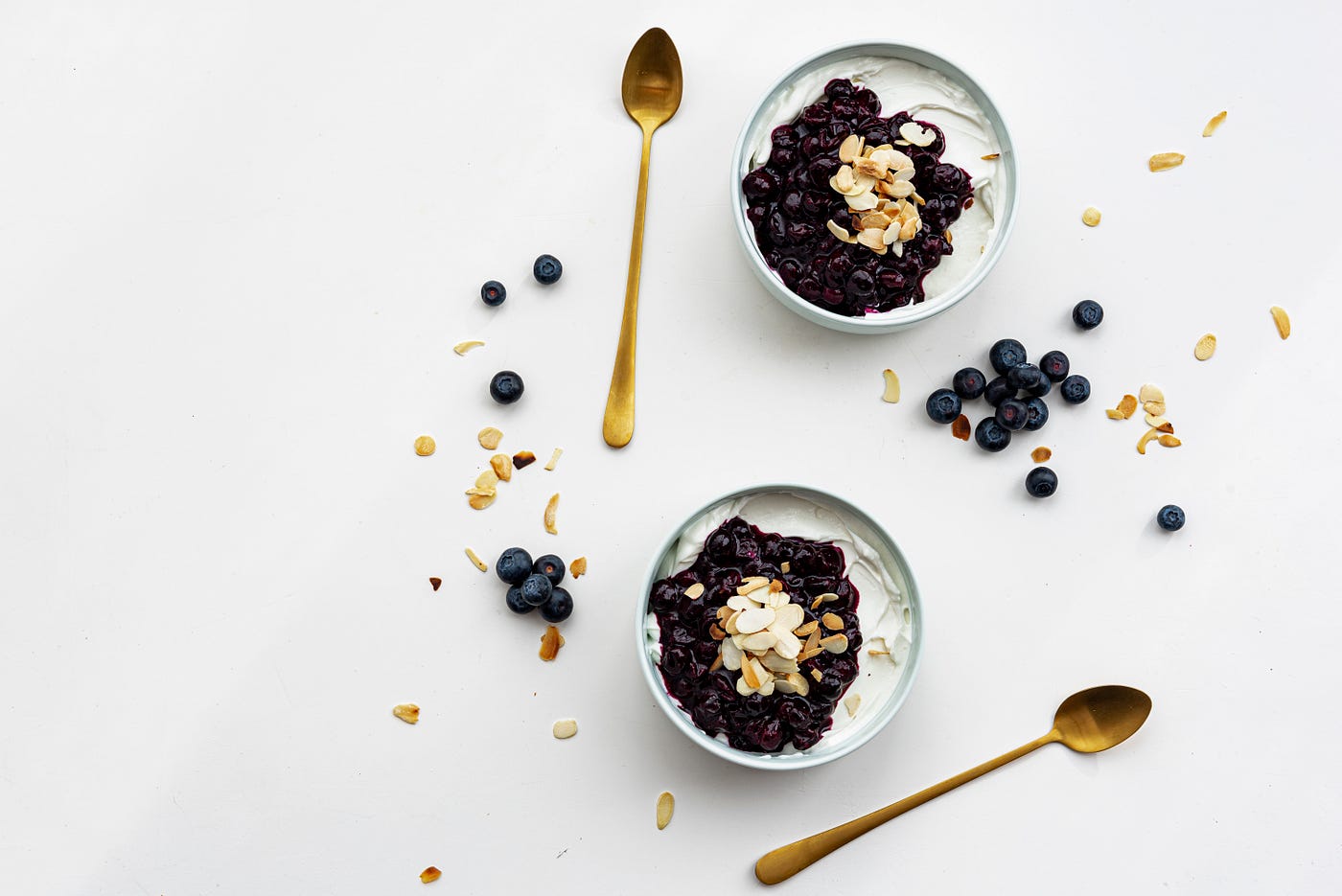 Two bowls of yogurt, topped by blueberries. There are berries and some cereal flakes scattered around the bowls. Two long gold-colored spoons sit on a white background.