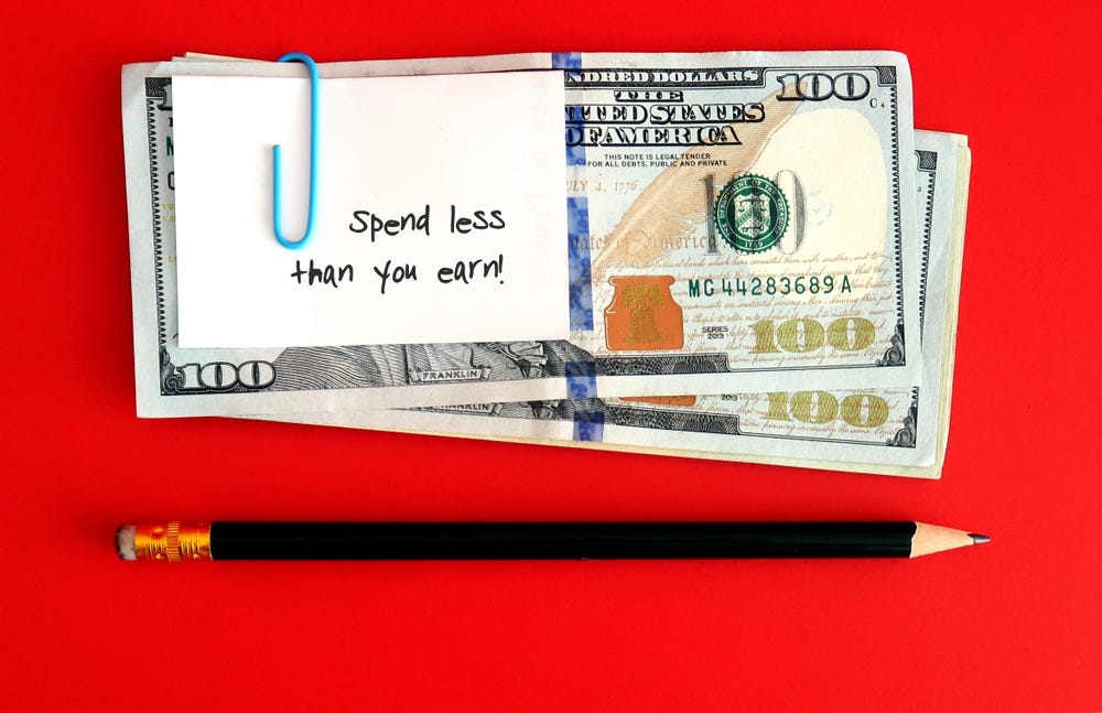 a bundle of dollars with a note that says 'spend less than you earn' and a pendil against a red background. The message encourages budgeting.
