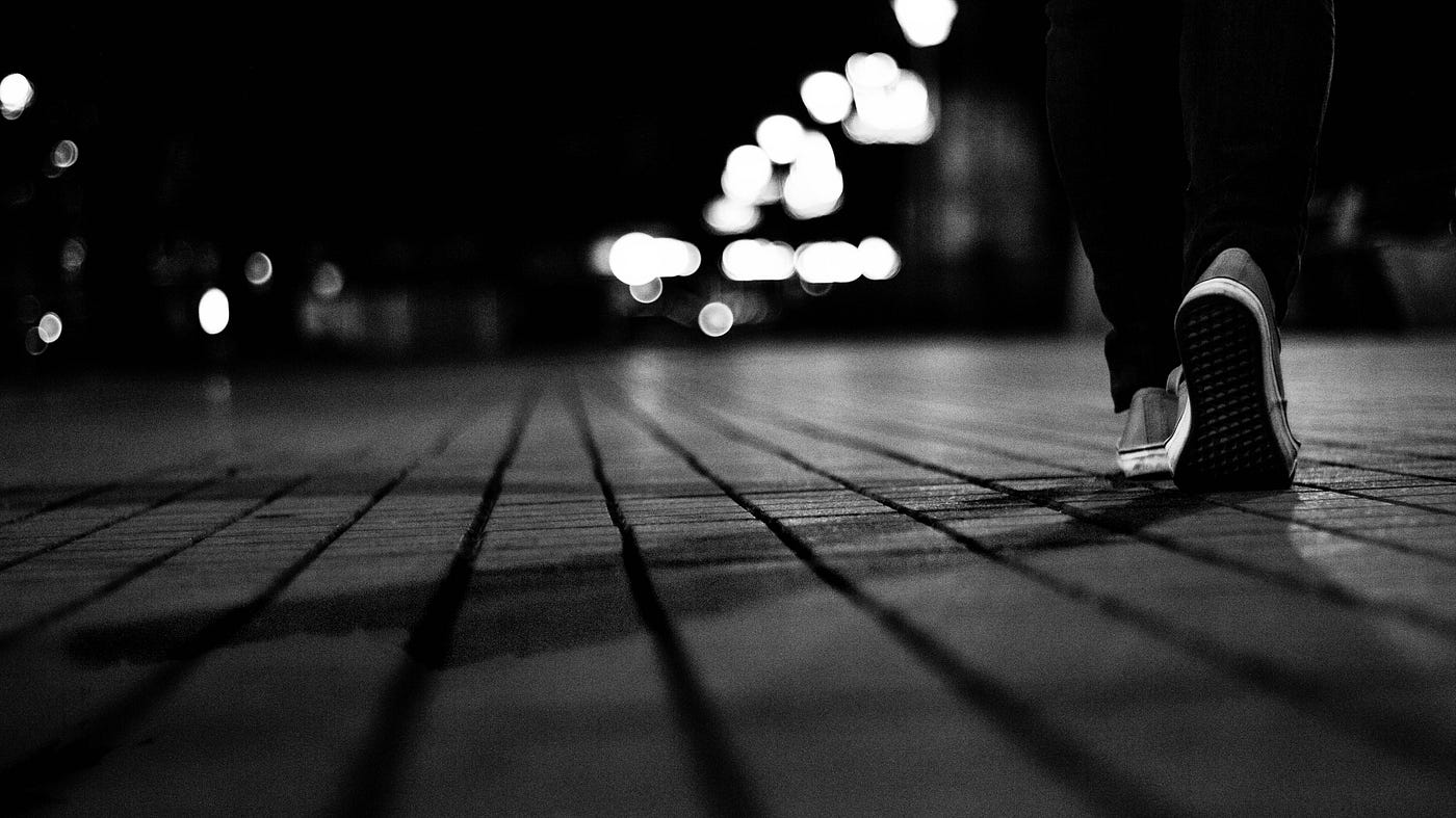 Black and white image of a person walking away from us. S/he is on the far right of the image, wears sneakers, and is seen from the knee level down. It is night and we see lights in the middle distance.