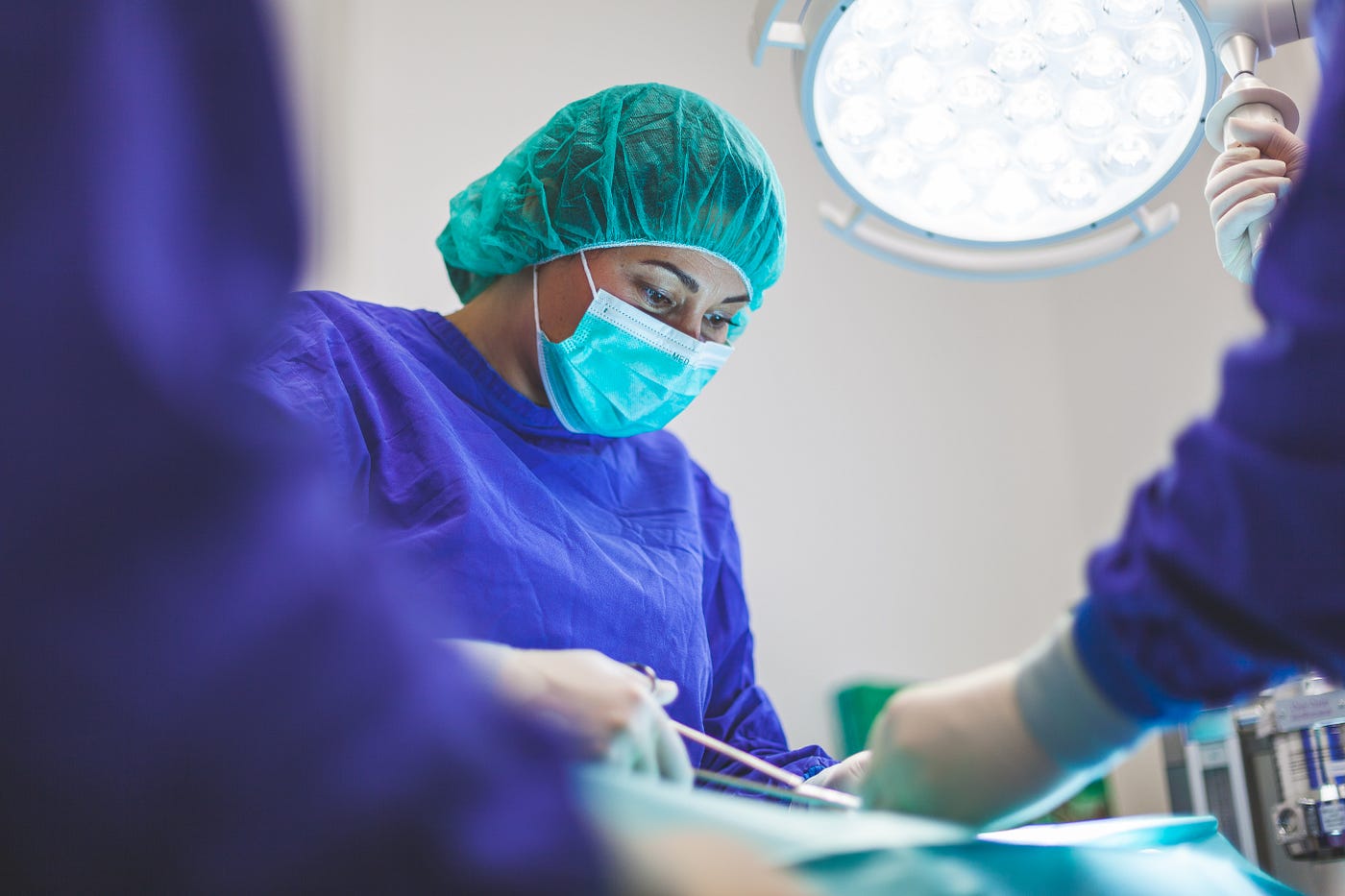 A female surgeon works in the operating room. She dons a green face mask and a green hair covering. New research suggests hospital-provided single-use undergarments improve the patient experience and satisfaction.